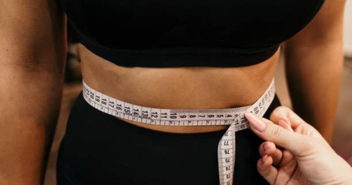 A person measuring their waist with a white measuring tape, wearing black athletic clothing.
