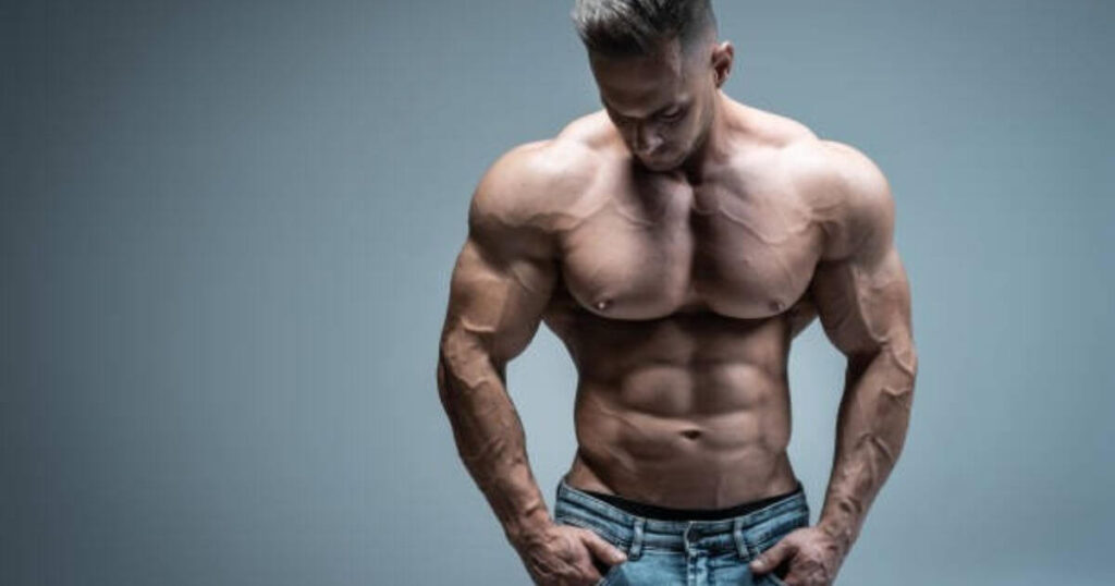 A muscular man with defined abs and arms, wearing jeans, standing against a neutral background
