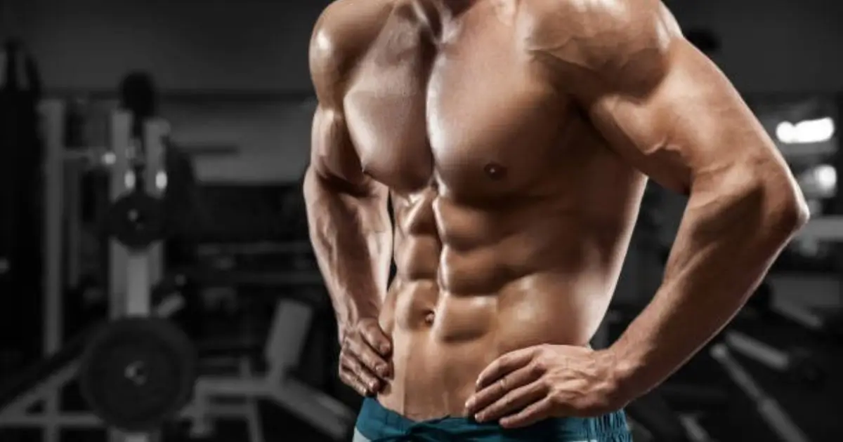 A muscular, shirtless individual with defined abs and arms stands confidently in a gym setting, showcasing their toned physique.