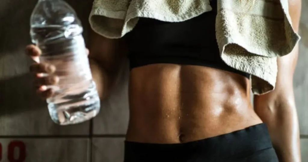 A person with a toned midsection is holding a clear water bottle, wearing a black sports bra and shorts, with a towel draped over their shoulders.
