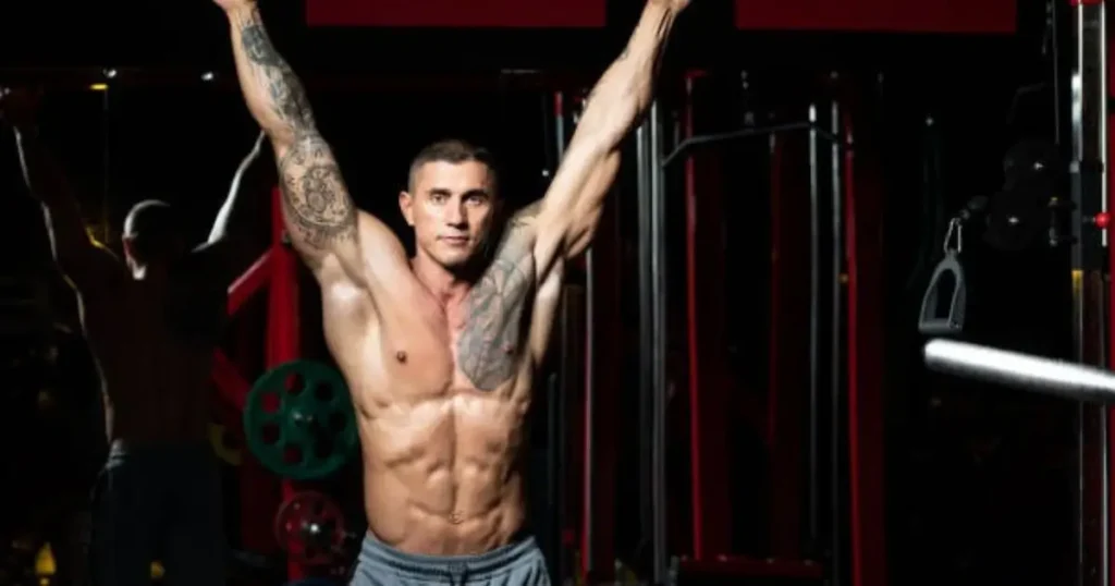 A muscular man with tattoos on his arms stands shirtless in a gym, raising both arms triumphantly.