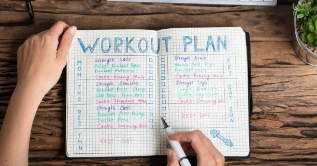 A person is holding a pen and marking off exercises on a detailed workout plan written in a grid notebook. The plan includes strength, cardio, and rest days for each day of the week.
