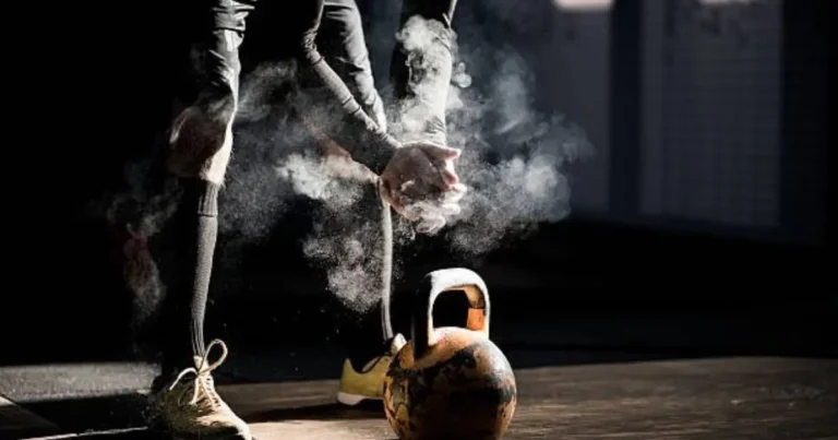 A person is applying chalk to their hands while preparing for a kettlebell workout. The chalk dust is visibly flying off as they rub it into their palms, emphasizing the focus and intensity of their preparation.