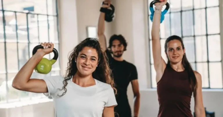 Three individuals are exercising in a bright, modern gym, each holding a kettlebell and smiling as they raise their arms.