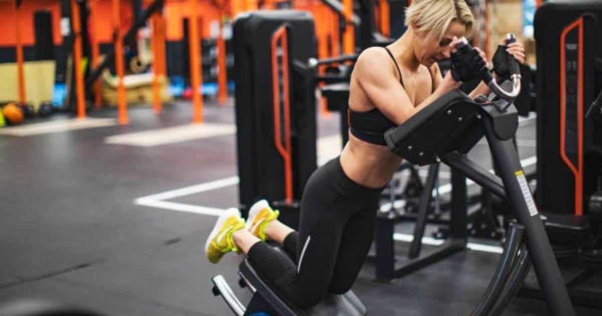 A woman is performing an exercise on a gym machine, focusing intently as she works out her upper body. She is wearing a black sports bra and leggings with yellow sneakers.