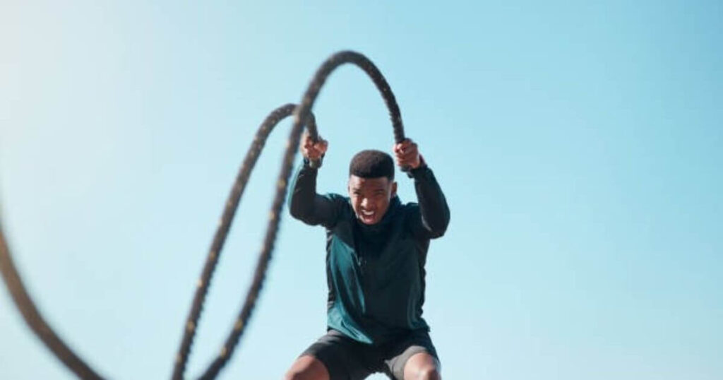 A person is vigorously swinging a battle rope, demonstrating intense physical effort against a clear blue sky backdrop.