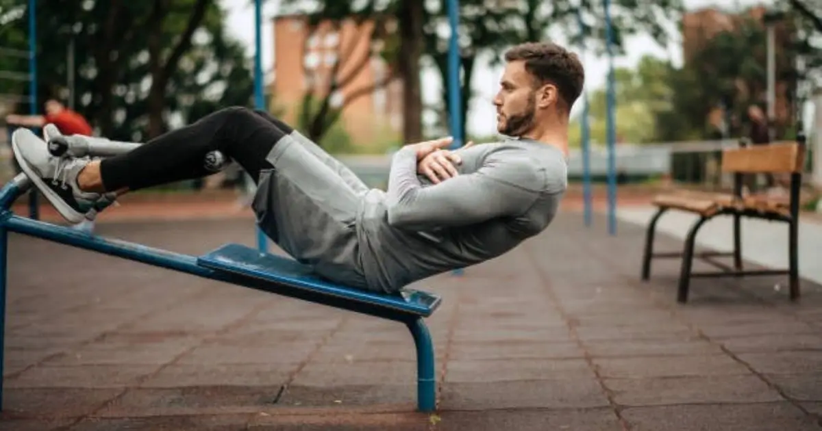 A man is performing an exercise on a blue outdoor bench, lying on his back with his legs raised and arms crossed over his chest.