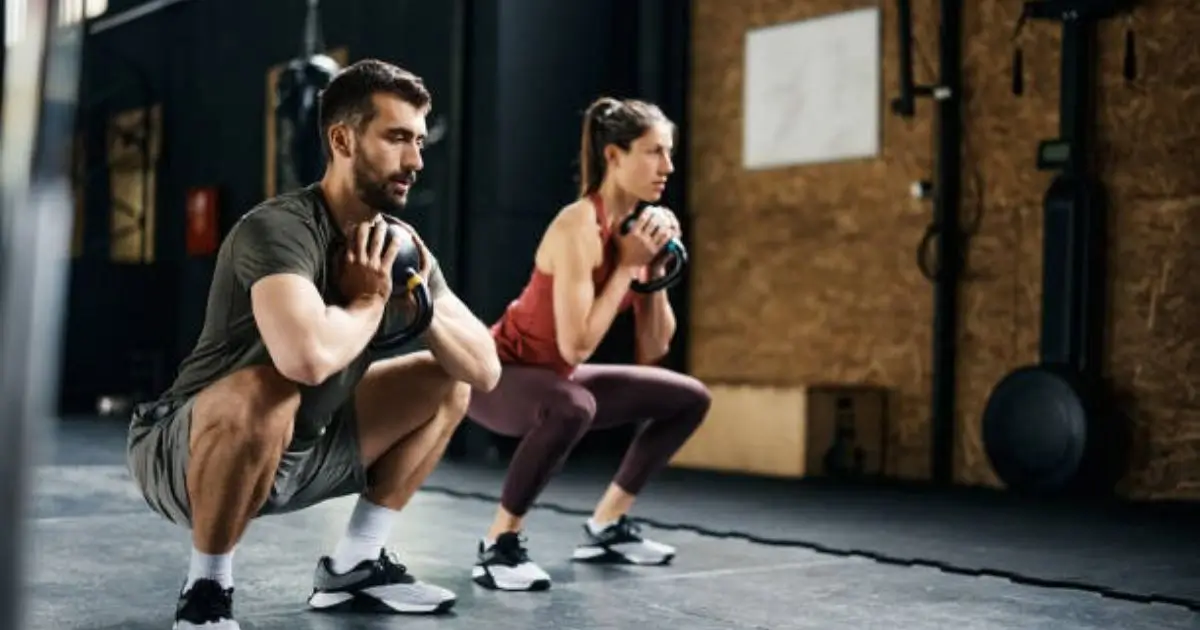 A man and a woman are performing kettlebell squats in a gym setting. Both individuals are holding kettlebells, with the man on the left and the woman on the right. They are dressed in athletic attire, focusing intently on their form.