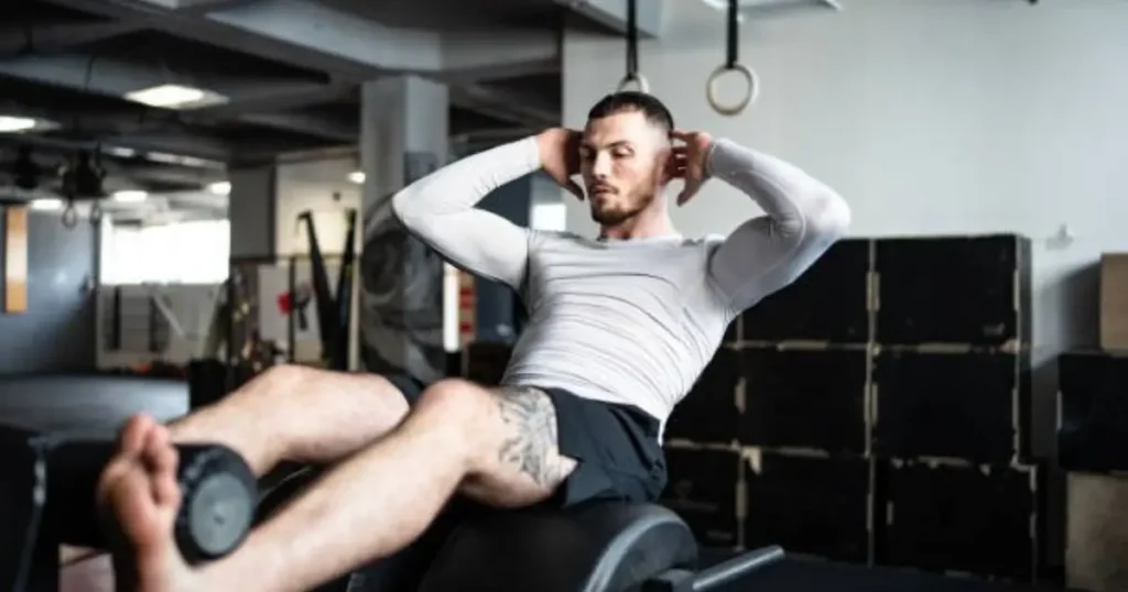 A man is performing an abdominal exercise on a weight bench in a gym, holding a weight plate between his feet.