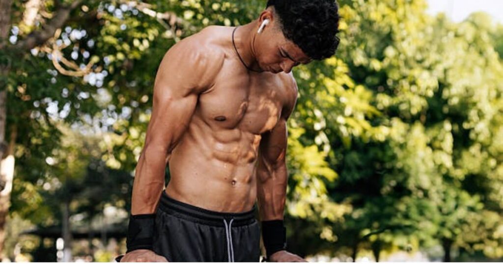 A muscular man is outdoors, shirtless, wearing black athletic shorts and wristbands. He has earbuds in his ears and is looking downward with a focused expression.