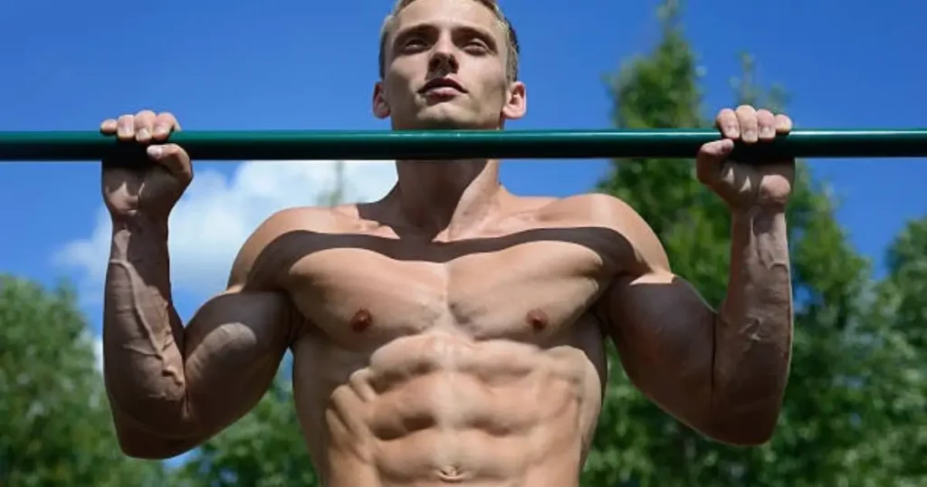 A muscular man is performing a pull-up on an outdoor green bar, showcasing his defined physique against a backdrop of trees and a clear blue sky.