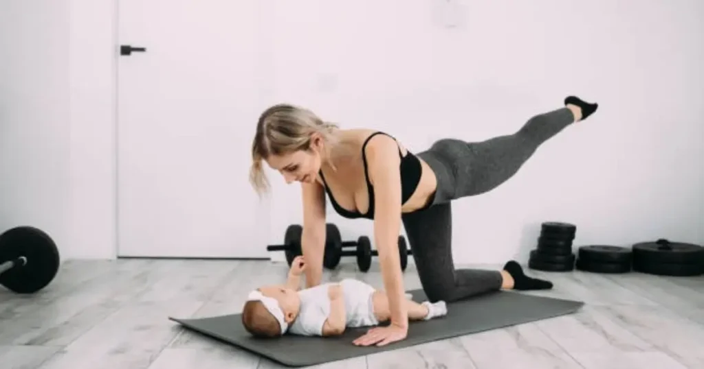 A woman is performing a side plank exercise on a yoga mat while interacting with a baby lying on the mat. She is wearing workout attire, and there are weights and dumbbells in the background.