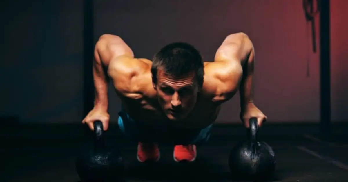 A muscular man is performing a push-up exercise using kettlebells as hand supports, demonstrating strength and focus in a dimly lit gym environment.