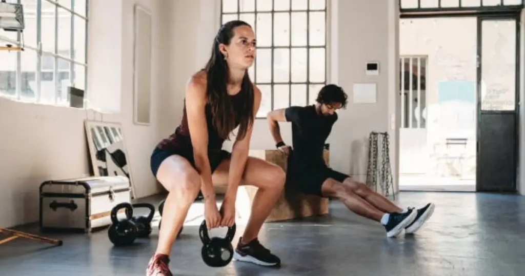 Two individuals are performing a workout in a bright, spacious gym. The person in the foreground is doing a kettlebell squat, while the person in the background is stretching or preparing for an exercise. The setting includes natural light streaming through large windows and fitness equipment like kettlebells and resistance bands.