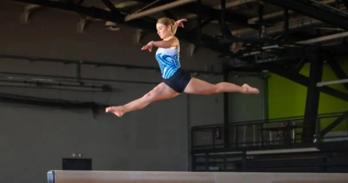 A gymnast in a blue and white leotard is performing a split leap on the balance beam, showcasing precision and grace in a gymnasium setting.