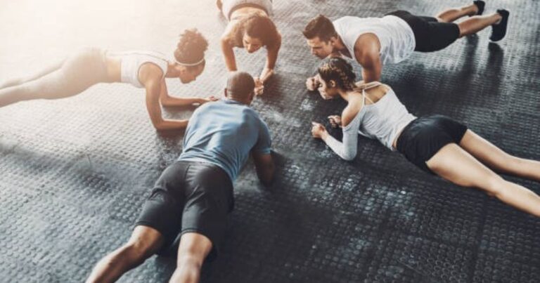 A group of five individuals is engaged in a synchronized workout on a gym floor, performing push-ups and plank exercises in unison.
