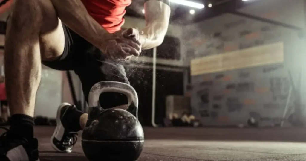 A person in a red shirt and black shorts is preparing to lift a kettlebell, with chalk dust visible as they grip the handle.