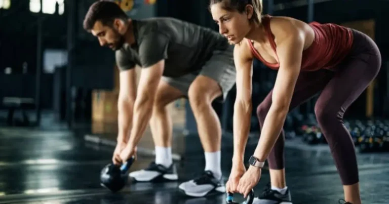 A man and a woman are performing kettlebell exercises in a gym, both bent over with their hands gripping the handles.