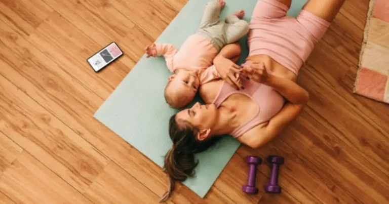 "A woman is lying on a yoga mat with her baby, holding dumbbells while exercising at home."