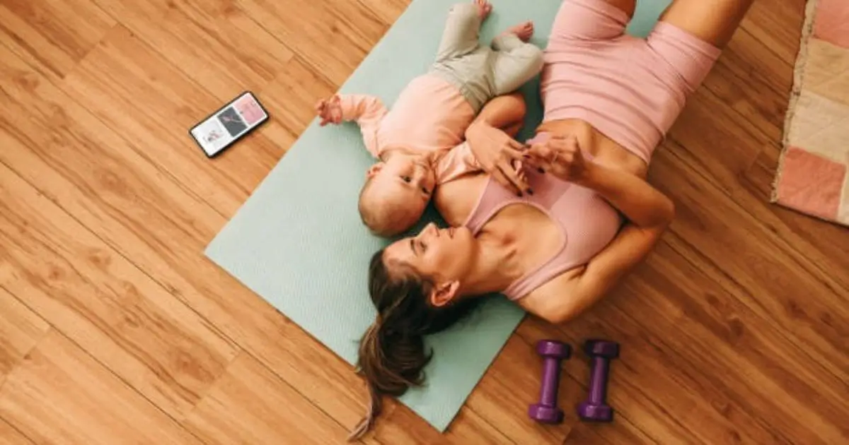 "A woman is lying on a yoga mat with her baby, holding dumbbells while exercising at home."