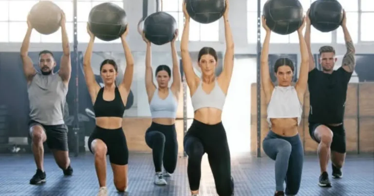 "A group of individuals performing a medicine ball exercise in a gym, holding the balls above their heads while in a squat position."