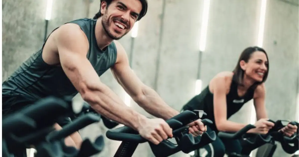"Two individuals are smiling while exercising on stationary bikes in a modern gym setting."