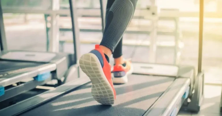 A close-up of a person's feet wearing bright orange and white sneakers, running on a treadmill in a gym setting.