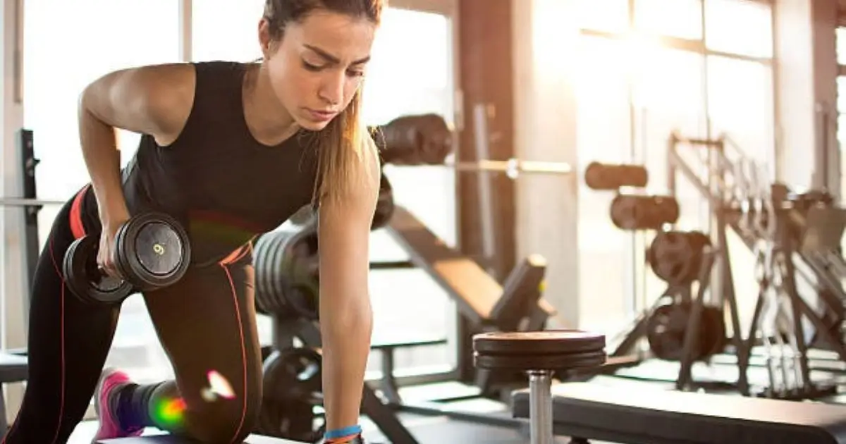 A woman in athletic attire performing a dumbbell row exercise at a gym, focusing intently on her form.