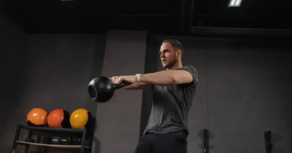 A man is performing a kettlebell exercise in a gym, holding a black kettlebell with both hands extended forward. The background features a rack of colorful kettlebells and a dark, minimalist setting.
