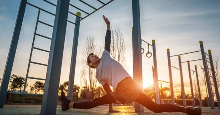 "A man performs a lunge stretch at an outdoor gym during sunset, surrounded by pull-up bars and exercise equipment, with warm golden light illuminating the scene."