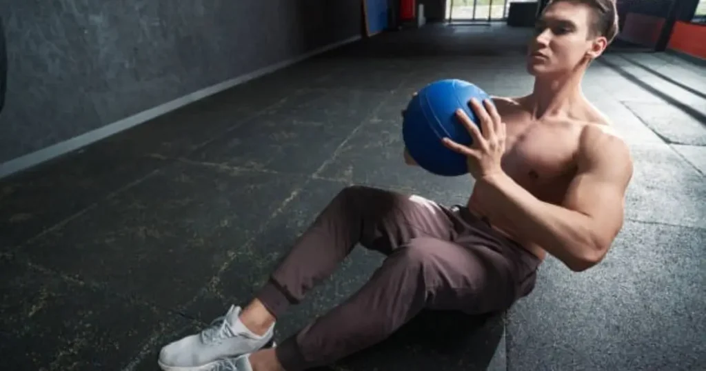 A muscular man is performing an exercise on the floor of a gym, holding a blue medicine ball against his chest while sitting with his legs bent.