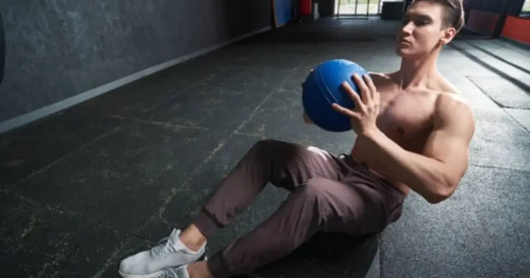 A muscular man is performing an exercise on the floor of a gym, holding a blue medicine ball against his chest while sitting with his legs bent.