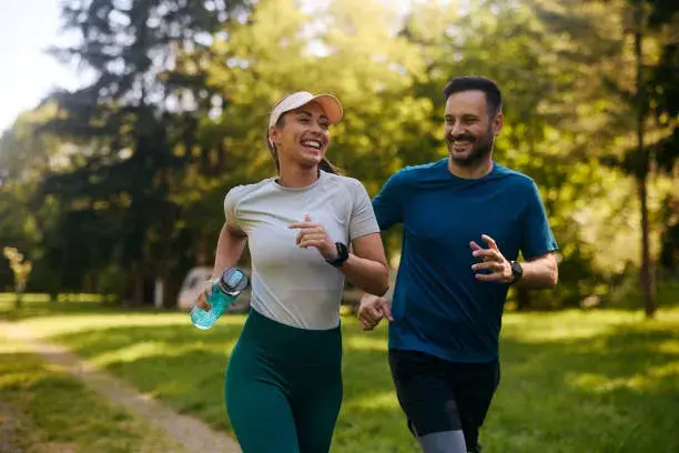 "Two individuals are jogging together in a lush, green park on a sunny day."