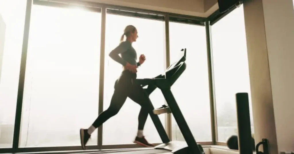 A woman is running on a treadmill in front of large windows, with sunlight streaming through the glass.