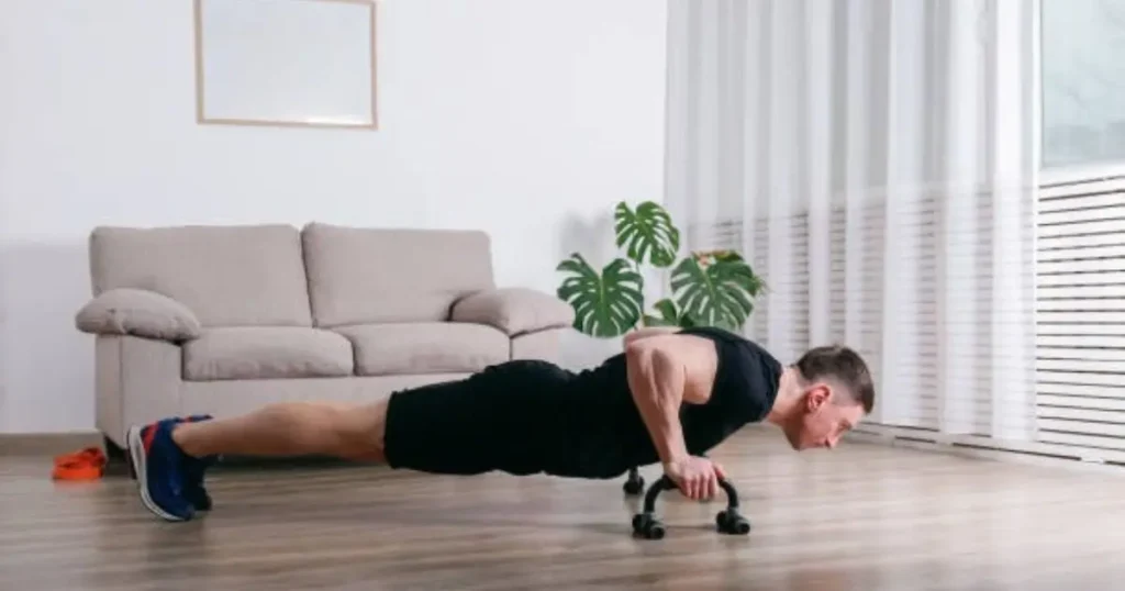 A man is performing push-ups at home, using a resistance band for added intensity. He is in a living room with a couch and a potted plant in the background.