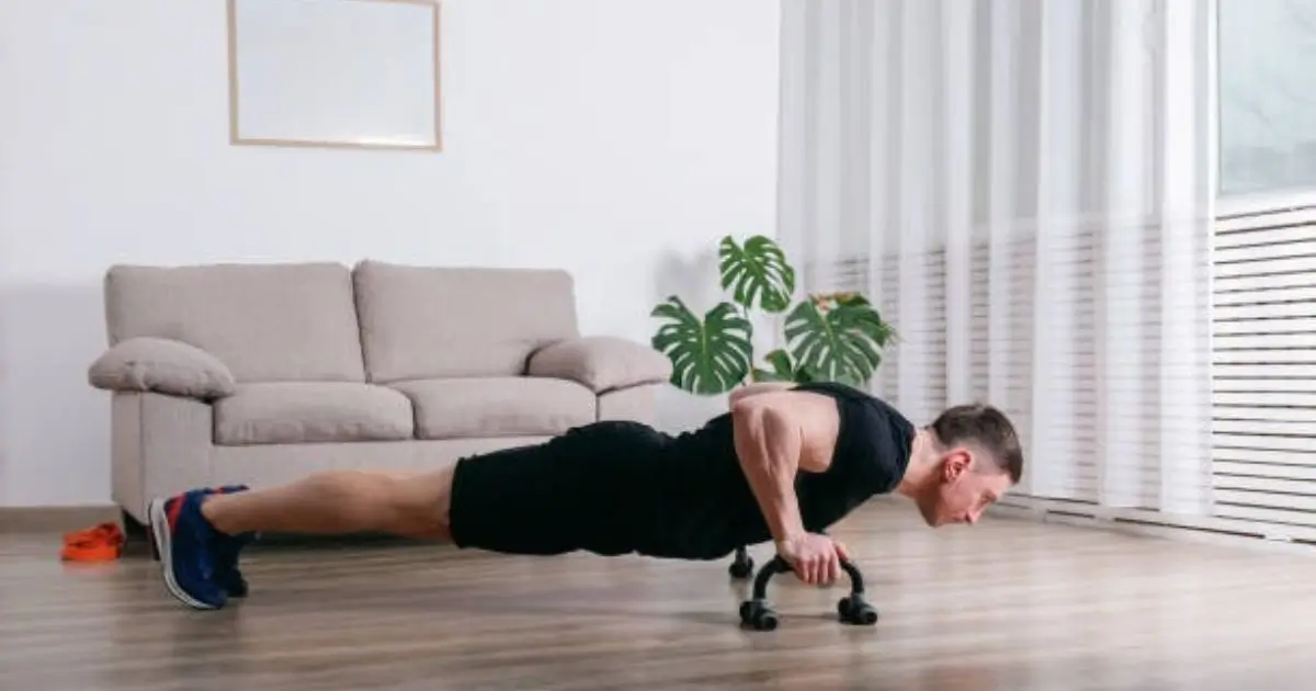 A man is performing push-ups at home, using a resistance band for added intensity. He is in a living room with a couch and a potted plant in the background.