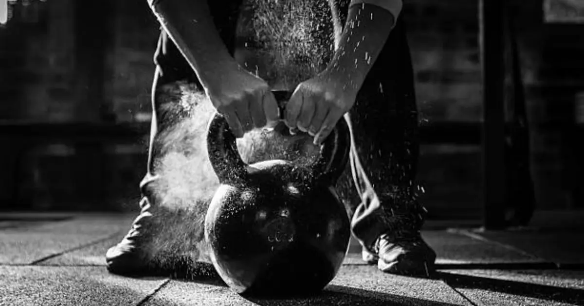 A person is gripping a kettlebell, with chalk dust visible as they prepare for a workout in a dimly lit gym setting.