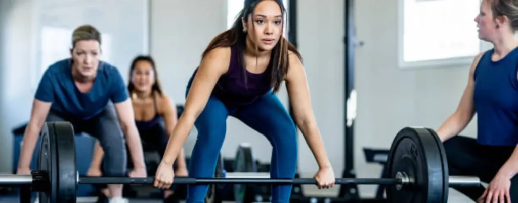 A group of women is engaged in a weightlifting workout, with one woman in the foreground performing a barbell exercise while others support and observe.