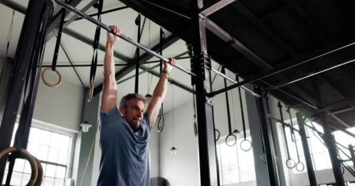 A man is performing a pull-up exercise on a horizontal bar in a well-lit, industrial-style gym with hanging rings and equipment visible in the background.